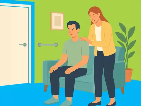 Eine Frau legt beruhigend die Hand auf die Schulter eines Mannes mit geistiger Behinderung, der auf einem Sofa in einem barrierefreien Wohnzimmer sitzt.