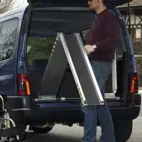 Mann lädt eine Klapprampe in den Kofferraum eines Busses, neben einem Rollstuhl. Kleidung: Dunkelblauer Pullover, Jeans.