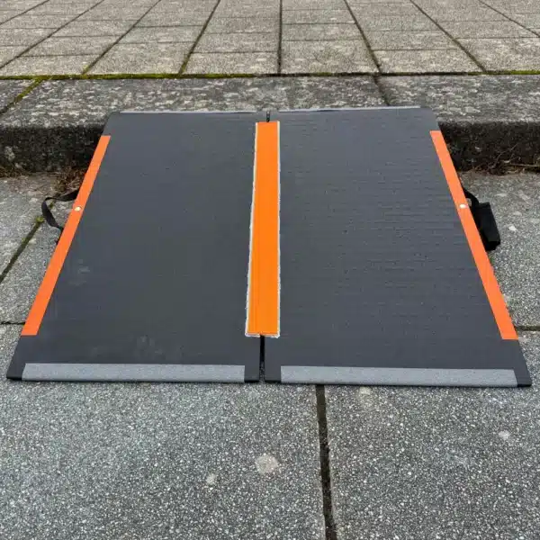 Faltbare Rampe aus rutschfestem Material mit orangefarbener Markierung, auf einem gepflasterten Boden platziert.