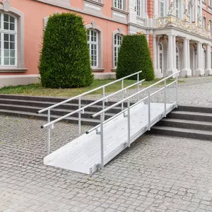 Eine Rollstuhlrampe aus Metall mit Geländer ist über einer Treppe vor einem historischen Gebäude mit großen Fenstern installiert.
