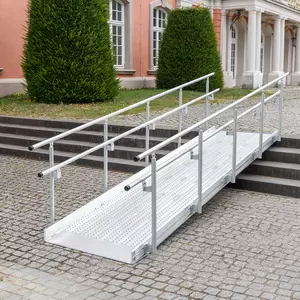 Vor einem Gebäude mit großen Fenstern und Grünflächen ist eine Rollstuhlrampe aus Metall mit Geländer über Steinstufen installiert.