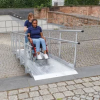 Zwei Frauen benutzen eine Rollstuhlrampe aus Aluinium im Freien; eine Frau schiebt die andere im Rollstuhl die Rampe hinauf.