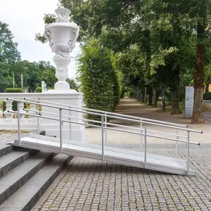 Eine Rampe für Rollstühle führt über Steinstufen im Freien neben einem von Bäumen und einer großen weißen Urne gesäumten Gehweg.