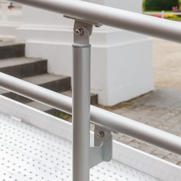 Nahaufnahme eines Metallhandlaufs und eines Stützpfostens an einer für Rollstuhlfahrer zugänglichen Rampe im Freien in der Nähe von Stufen und einem Gehweg.