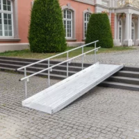 Vor einem historischen Gebäude mit großen Fenstern und Sträuchern ist eine Rollstuhlrampe aus Metall mit Handläufen über Steinstufen angebracht.