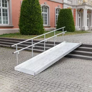 Vor einem historischen Gebäude mit großen Fenstern und Sträuchern ist eine Rollstuhlrampe aus Metall mit Handläufen über Steinstufen angebracht.