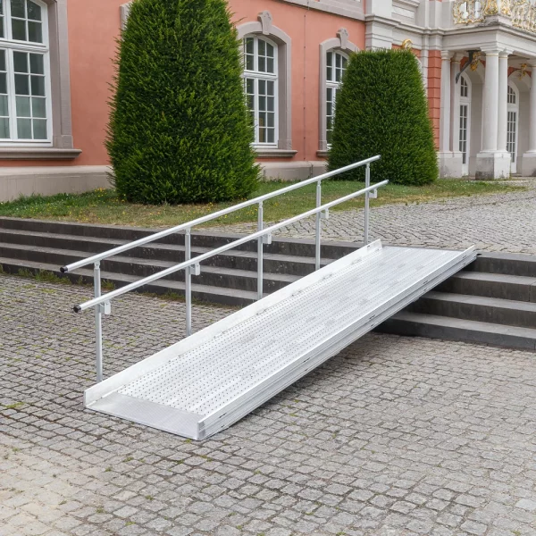 02 Geländer einseitig für Rollstuhlrampe fest LINKS diagonal rechts Vor einem historischen Gebäude mit großen Fenstern und Sträuchern ist eine Rollstuhlrampe aus Metall mit Handläufen über Steinstufen angebracht.