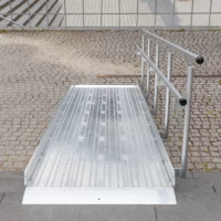 Eine Rollstuhlrampe aus Metall mit erhöhten Rändern und Handläufen wird über Steinpflaster und Stufen installiert.
