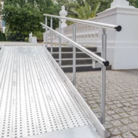 Eine Rollstuhlrampe aus Aluminium mit Handläufen führt zu Steinstufen vor einem weißen Gebäude mit Grün im Hintergrund.