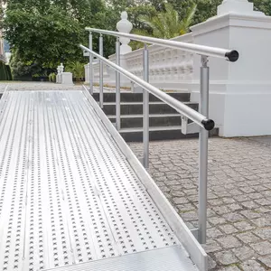 Eine Rollstuhlrampe aus Aluminium mit Handläufen führt zu Steinstufen vor einem weißen Gebäude mit Grün im Hintergrund.
