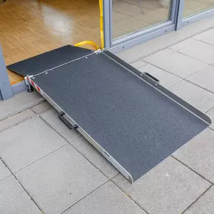Eine tragbare Rollstuhlrampe ist an einer Türschwelle angebracht und überbrückt den Boden im Innenbereich auf die Steinplatten im Freien.