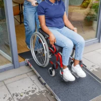 Eine Person im Rollstuhl wird von einer anderen Person über eine tragbare Rampe an einer Türöffnung geschoben.