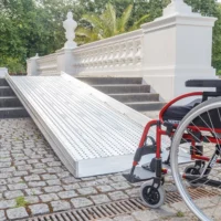 Eine Rampe für Rollstühle führt neben einem weißen Geländer eine Treppe hinauf; am unteren Ende der Rampe befindet sich ein Rollstuhl.