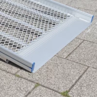 Eine Rollstuhlrampe aus Metall mit einer geriffelten Oberfläche ruht auf grauen Pflastersteinen im Freien.