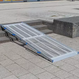 Eine tragbare Rollstuhlrampe aus Aluminium ist über drei Stufen auf einem gepflasterten Außengelände angebracht.
