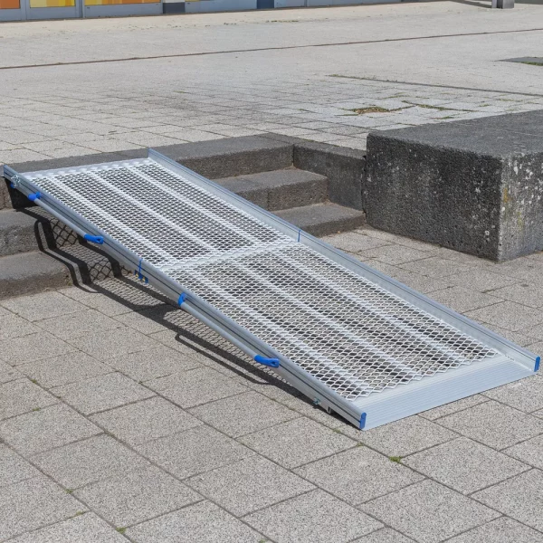 10 Rollstuhlrampe Flächenrampe klappbar diagonal links Eine tragbare Rollstuhlrampe aus Aluminium ist über drei Stufen auf einem gepflasterten Außengelände angebracht.