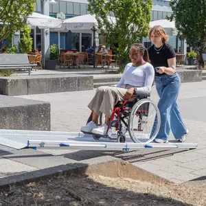 Eine Frau schiebt eine andere Frau im Rollstuhl auf eine tragbare Rollstuhlrampe auf einem öffentlichen Platz in der Nähe der Sitzgelegenheiten eines Straßencafés.
