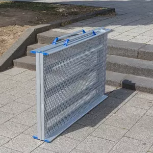 Eine tragbare Rollstuhlrampe aus Aluminium steht zusammengeklappt neben mehreren Betonstufen in einem Außenbereich.