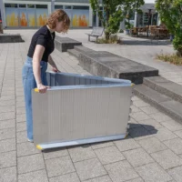 Eine Person klappt eine faltbare Rollstuhlrampe auf einem gepflasterten Außengelände in der Nähe von Stufen und Bänken auf.