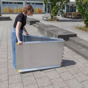 Eine Person klappt eine faltbare Rollstuhlrampe auf einem gepflasterten Außengelände in der Nähe von Stufen und Bänken auf.