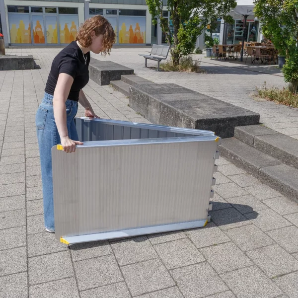 Eine Person klappt eine faltbare Rollstuhlrampe auf einem gepflasterten Außengelände in der Nähe von Stufen und Bänken auf.