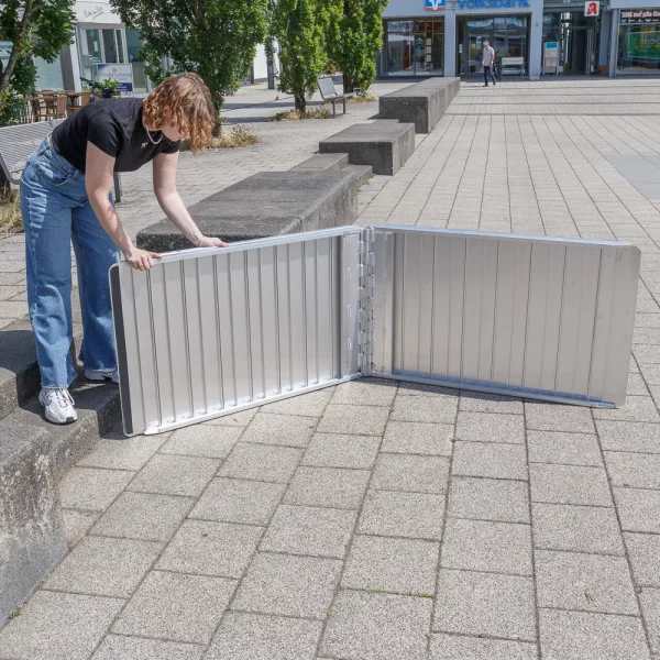 Eine Person klappt eine faltbare Rollstuhlrampe auf einem gepflasterten Außengelände mit Bänken und Bäumen in der Nähe auseinander.
