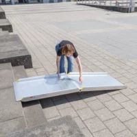 Eine Person legt eine tragbare Aluminium-Rollstuhlrampe auf Betonstufen im Freien auf.