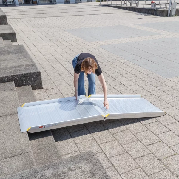 Eine Person legt eine tragbare Aluminium-Rollstuhlrampe auf Betonstufen im Freien auf.
