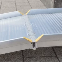Nahaufnahme des Klappmechnismus einer Aluminium-Rollstuhlrampe mit gelben Eckschützern auf einer gepflasterten Außenfläche.