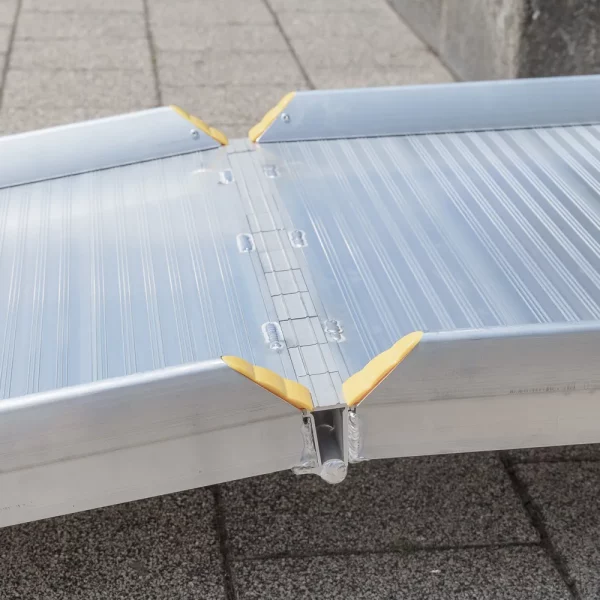 Nahaufnahme des Klappmechnismus einer Aluminium-Rollstuhlrampe mit gelben Eckschützern auf einer gepflasterten Außenfläche.