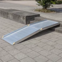 Eine klappbare Rollstuhlrampe liegt auf Stufen und auf einem gepflasterten Gehweg im Freien und ermöglicht Rollstuhlfahrern den Zugang.
