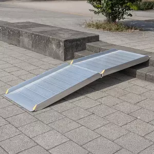 Eine klappbare Rollstuhlrampe liegt auf Stufen und auf einem gepflasterten Gehweg im Freien und ermöglicht Rollstuhlfahrern den Zugang.