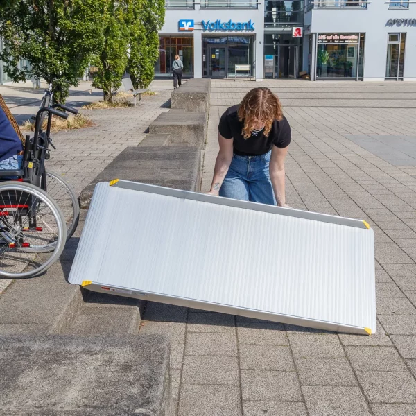 Eine Person legt eine tragbare Rollstuhlrampe auf eine Treppe aus 2 Stufen in der Nähe eines Rollstuhls im Freien in einem städtischen Gebiet auf.
