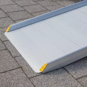 Nahaufnahme des unteren Endes einer tragbaren Rollstuhlrampe aus Aluminium mit gelben Eckschutzvorrichtungen auf einer gepflasterten Steinfläche.