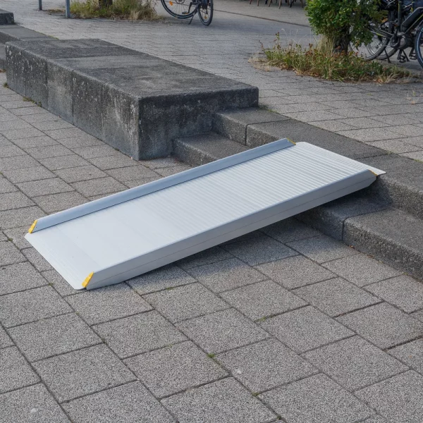 Eine tragbare Aluminium-Rollstuhlrampe liegt auf einer Treppe mit 2 Stufen um den barrierefreien Zugang vom gepflasterten Platz auf den höher liegenden Gehweg zu ermöglichen.
