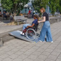 Eine Frau schiebt eine andere Frau im Rollstuhl auf einer tragbare Aluminium-Rollstuhlrampe über eine Treppe mit 2 Stufen in der Nähe von Sitzgelegenheiten im Freien und Bäumen.