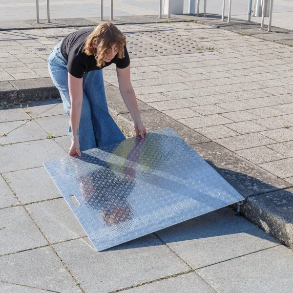Eine Person positioniert eine Rollstuhlrampe aus Aluminium über einen Bordstein auf einem gepflasterten Außengelände.