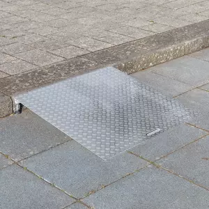 Eine Rollstuhlrampe aus Aluminium liegt an einer einzelnen Betonstufe zwischen einem gepflasterten Gehweg und einer gepflasterten Fläche.