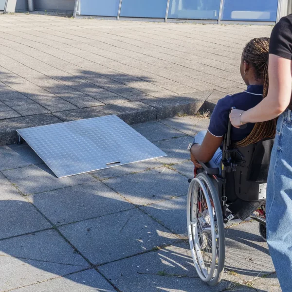 14 Rollstuhlrampe Keilbrücke mit Rollstuhl 3 Eine Person im Rollstuhl wird von einer Hilfsperson zu einer tragbaren Rollstuhlrampe aus Aluminium geschoben