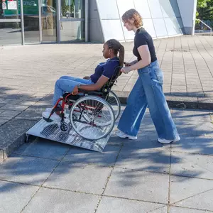 Eine Frau schiebt eine andere Frau im Rollstuhl über eine Rollstuhlrampe aus Aluminium auf den Bürgersteig vor einem modernen Gebäude.