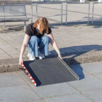 Eine Person richtet eine tragbare Aluminium-Rollstuhlrampe vor einer einzelnen Stufe im Freien auf einer gepflasterten Fläche aus.