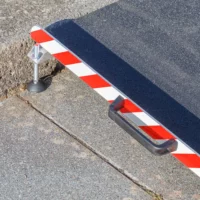 Nahaufnahme des linken Griffs einer Aluminium-Rollstuhlrampe mit rot- weißen Seitenstreifen. Aufgestellt vor einer einzelnen Stufe auf einer gepflasterten Fläche.