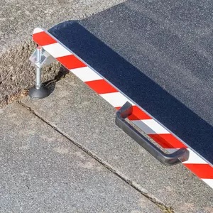 Nahaufnahme des linken Griffs einer Aluminium-Rollstuhlrampe mit rot- weißen Seitenstreifen. Aufgestellt vor einer einzelnen Stufe auf einer gepflasterten Fläche.
