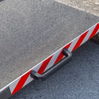 Nahaufnahme des rechten Griffs einer Aluminium-Rollstuhlrampe mit rot- weißen Seitenstreifen. Aufgestellt vor einer einzelnen Stufe auf einer gepflasterten Fläche.