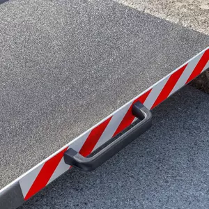 Nahaufnahme des rechten Griffs einer Aluminium-Rollstuhlrampe mit rot- weißen Seitenstreifen. Aufgestellt vor einer einzelnen Stufe auf einer gepflasterten Fläche.