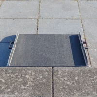 Eine mobile Aluminium-Rollstuhlrampe mit Griffen und rutschfester Korund Oberfläche steht vor einer einzelnen Stufe auf einer gepflasterten Ebene.