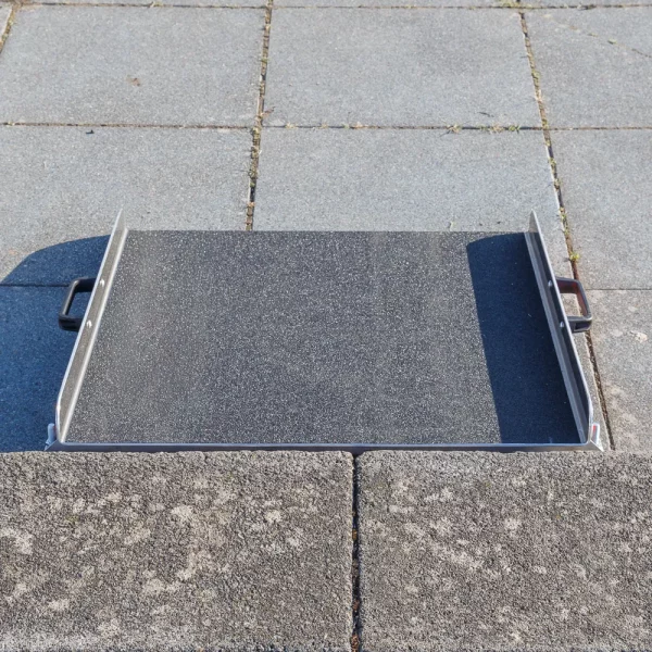 Eine mobile Aluminium-Rollstuhlrampe mit Griffen und rutschfester Korund Oberfläche steht vor einer einzelnen Stufe auf einer gepflasterten Ebene.