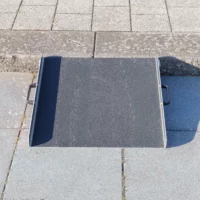 Eine mobile Aluminium-Rollstuhlrampe mit Griffen und rutschfester Korund Oberfläche steht gerade vor einer einzelnen Stufe auf einer gepflasterten Ebene.