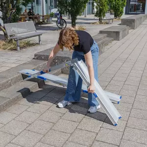 Eine Person klappt eine tragbare Rollstuhlrampe auf einem gepflasterten Außengelände in der Nähe von Bänken und Gebäuden aus.