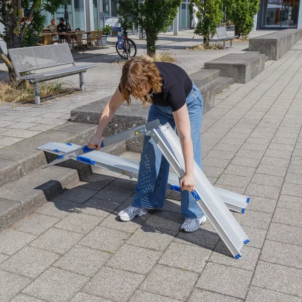 Eine Person klappt eine tragbare Rollstuhlrampe auf einem gepflasterten Außengelände in der Nähe von Bänken und Gebäuden aus.
